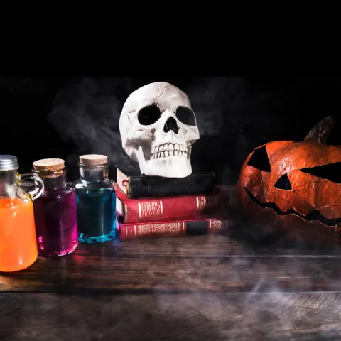 A skull rests on a pile of books next to three brightly colored potions jars.