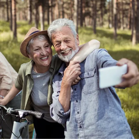 An adult woman balances on her bike with her arm thrown around a man holding a phone. The man is taking a picture of them both.