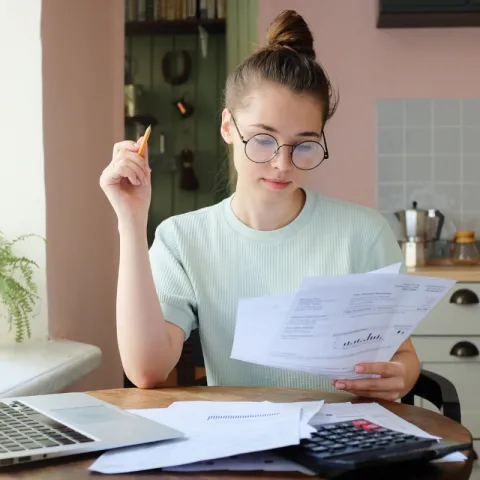A young woman looks thoughtfully at documents while holding a pen in the air.