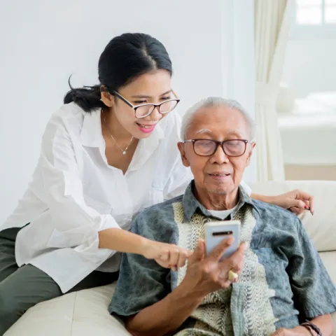 An older man sits in a chair looking at his phone. A younger woman looks over his shoulder, pointing at something on the device. Both are smiling.