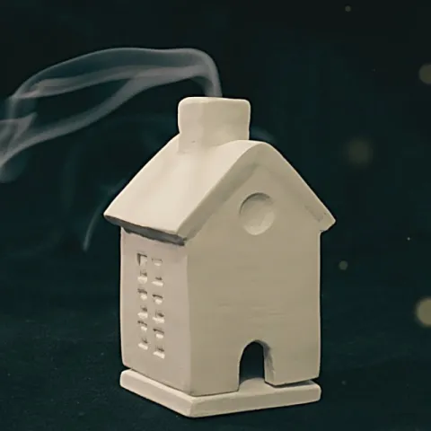 A small white house made of clay has a trail of incense smoke coming from the chimney.
