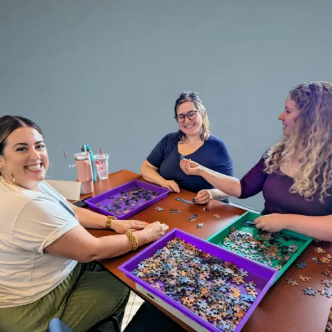 Three women sort puzzle pieces while smiling at the camera and each other.