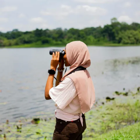A young black woman in hijab gazes through binoculars into the sky.