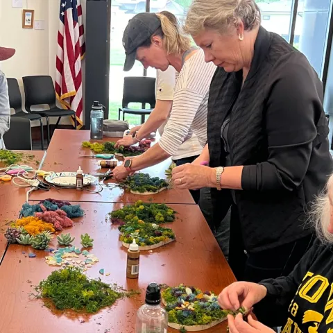 Several women stand around a table. They are gluing moss to wooden blocks.