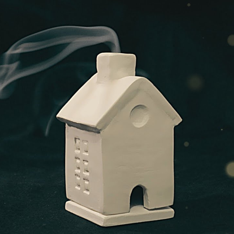 A small white house made of clay has a trail of incense smoke coming from the chimney.